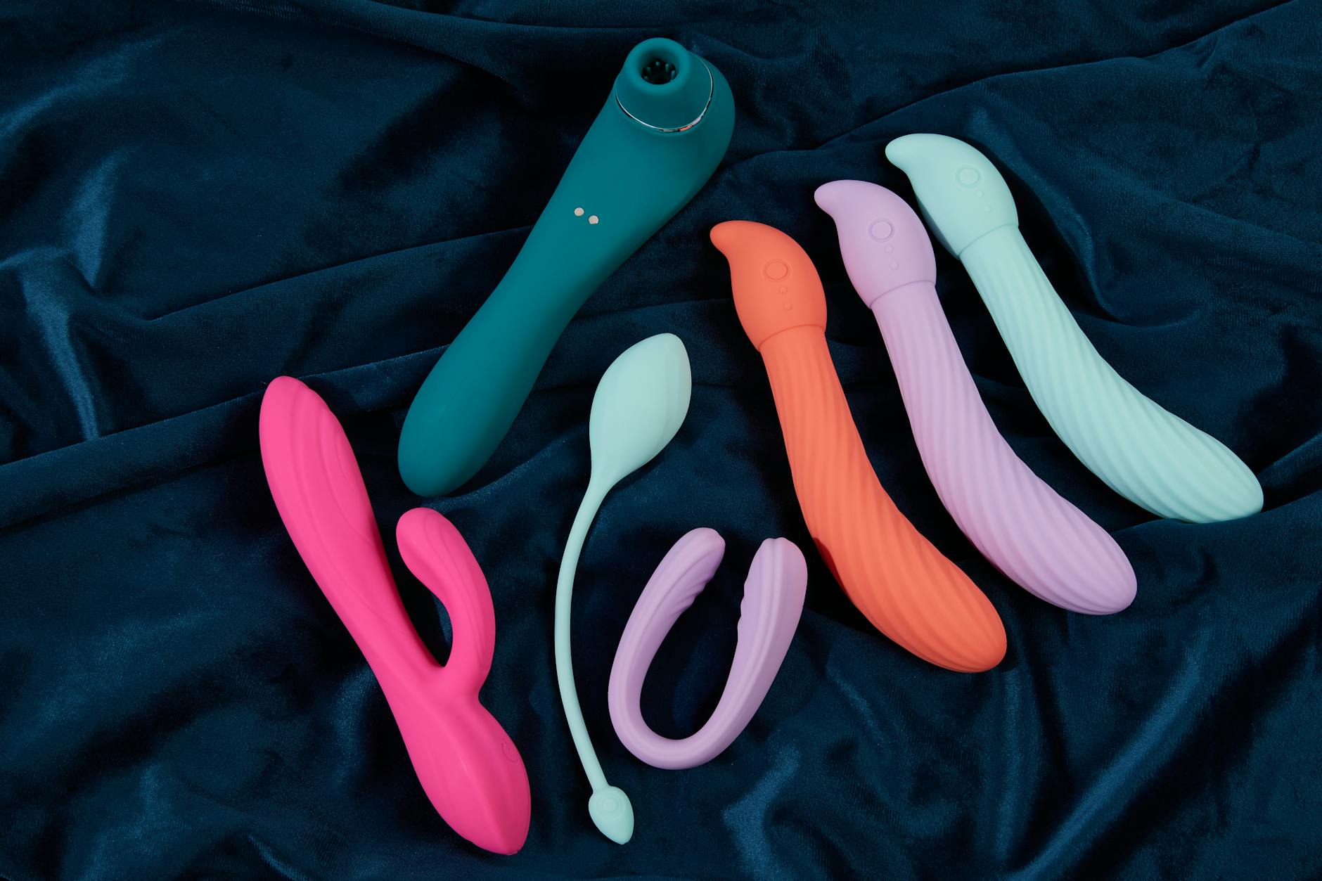 Array of colorful silicone clitoral vibrators displayed on white fabric, showing texture and design variation