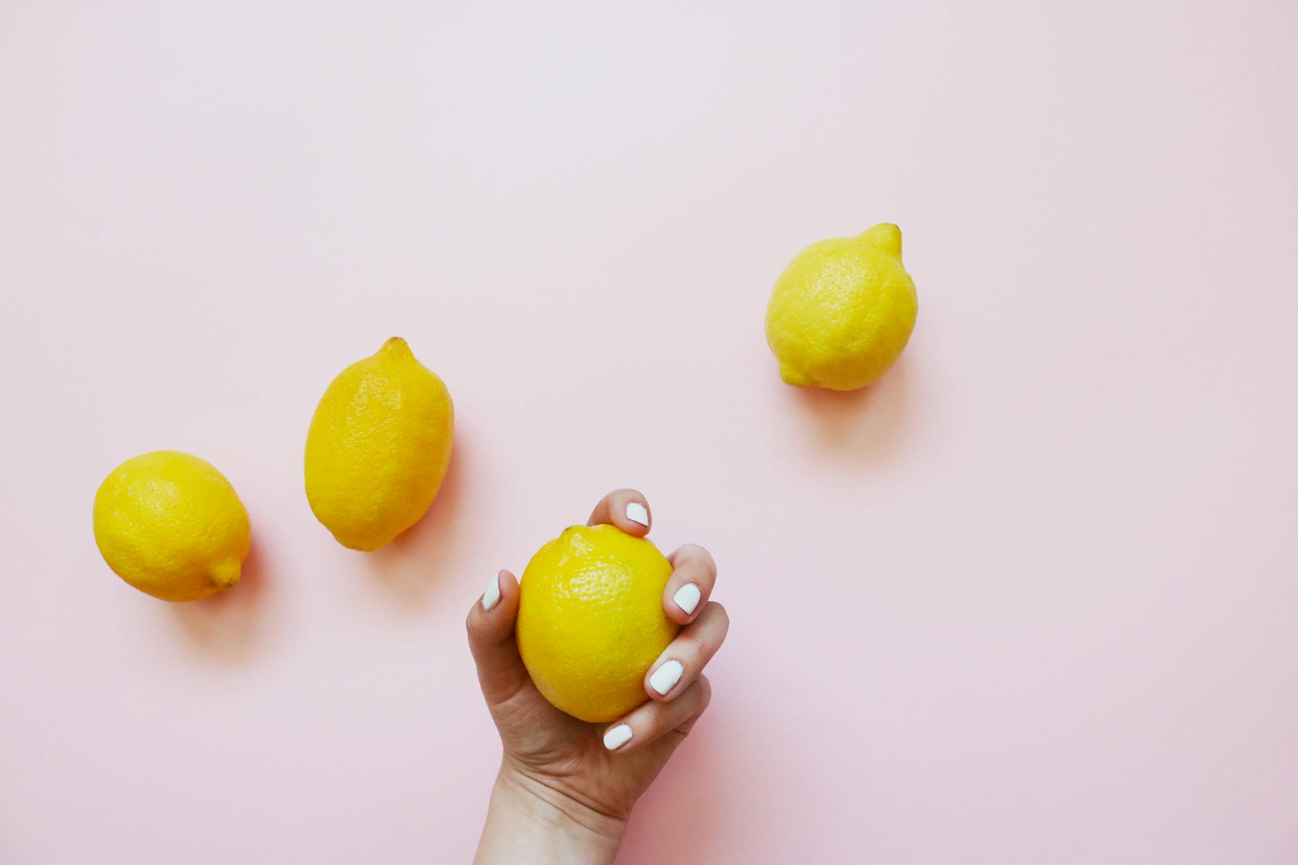 A hand holding a bright yellow lemon on a soft pink background, surrounded by additional lemons, representing the Lemon clitoral vibrator