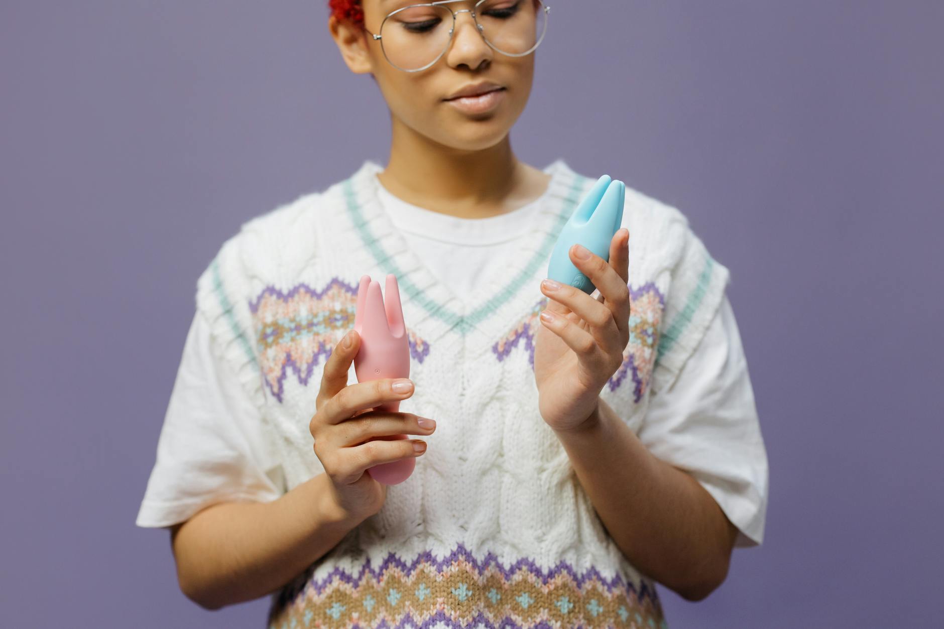Woman with eyeglasses holding blue and pink silicone vibrators in a contemplative manner